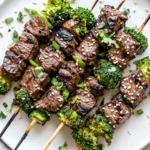 Grilled Beef And Broccoli Skewers On A White Plate Garnished With Fresh Herbs And Sesame Seeds