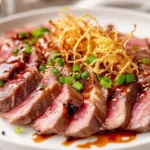Sliced Beef Tataki On A White Plate Drizzled With Yuzu Sauce, Garnished With Potato Straws And Green Onions