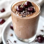 Cherry Mocha Smoothie In A Clear Glass Topped With Almond Butter And Cacao Nibs, Served On A White Plate