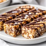 Close-Up Of Chewy Snack Bars Drizzled With Dark Chocolate On A White Plate, Garnished With Sea Salt.