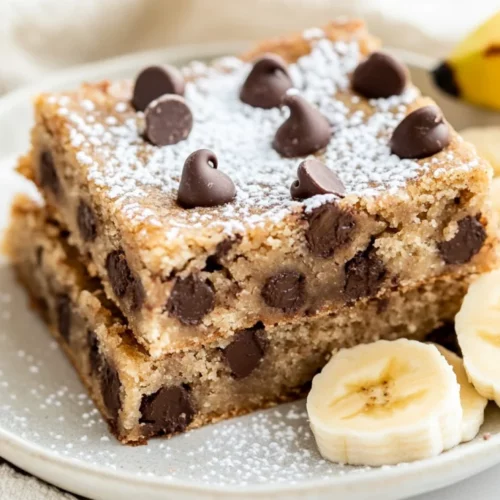 Gooey Chocolate Chip Banana Squares: The Best Banana Dessert Bars 2 Gooey Chocolate Chip Banana Squares On A White Plate With Powdered Sugar And Fresh Banana Slices