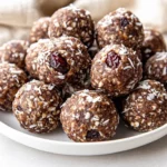 Plate Of Chocolate Date Energy Balls Dusted With Coconut, Arranged On A White Plate With Chopped Nuts And Cherries