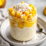 Coconut Mango Overnight Oats In A Glass Jar Topped With Fresh Mango And Shredded Coconut, Served With A Spoon