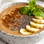 Coffee Smoothie Bowl Topped With Bananas, Chia Seeds, And Cocoa Powder Served In A White Bowl On A Neutral Surface