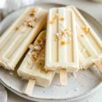 Creamy Coconut Popsicles On A White Plate, Topped With Coconut Flakes And Set Against A Neutral Background.