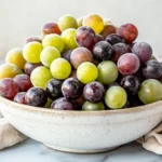 Bowl Of Frozen Grapes In A White Ceramic Bowl On A Marble Surface, Showcasing Vibrant Textures And Colors.