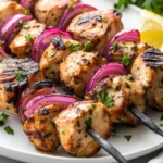 Grilled Chicken Kebabs On A White Plate Garnished With Parsley And Lemon Wedges, Showcasing Vibrant Colors.
