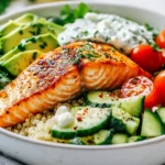 Vibrant Mediterranean Salmon Bowl With Flaky Salmon, Quinoa, And Fresh Veggies In A White Bowl, Topped With Tzatziki And Feta.