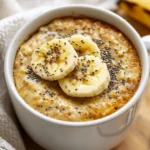 Warm Microwave Banana Oat Mug Cake Topped With Banana Slices And Chia Seeds, Served In A White Mug On A Wooden Surface