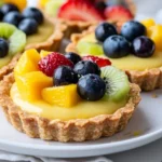 No-Bake Lemon Tarts With Fruit Topped With Fresh Strawberries, Kiwi, Blueberries, And Mango On A White Plate