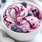 Bowl Of No Churn Blueberry Mascarpone Ice Cream Topped With Blueberry Jam And Fresh Blueberries, Served With A Gray Kitchen Towel