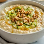 Bowl Of Overnight Oats With Zucchini Topped With Chopped Walnuts And Maple Syrup, Against A Neutral Background.