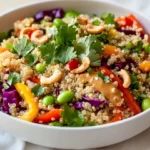 A Colorful Quinoa Vegetable Salad In A White Bowl Drizzled With Creamy Peanut Dressing And Topped With Cashews.