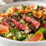 Steak Grapefruit Salad In A White Bowl, Topped With Fresh Herbs And Crispy Onions, Against A Neutral Background.