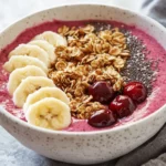 Tart Cherry Smoothie Bowl Topped With Banana Slices, Granola, And Chia Seeds In A White Bowl