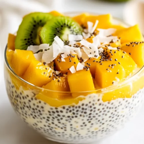 Bowl Of Tropical Chia Pudding Topped With Sliced Kiwi, Cubed Mango, And Toasted Coconut, Served With A Spoon.